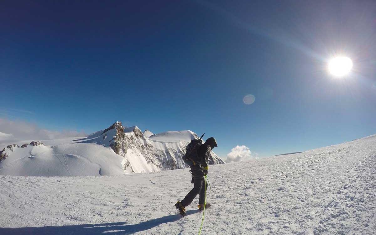 Alpinista cammina sulla neve con montagna innevata e cielo sereno sullo sfondo.