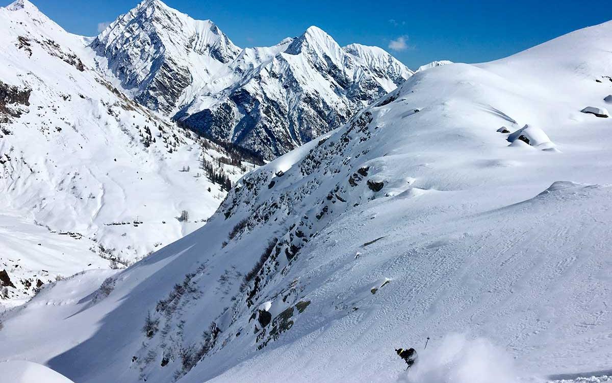 Scenari montani innevati con un sciatore in discesa, cielo limpido e montagne imponenti.