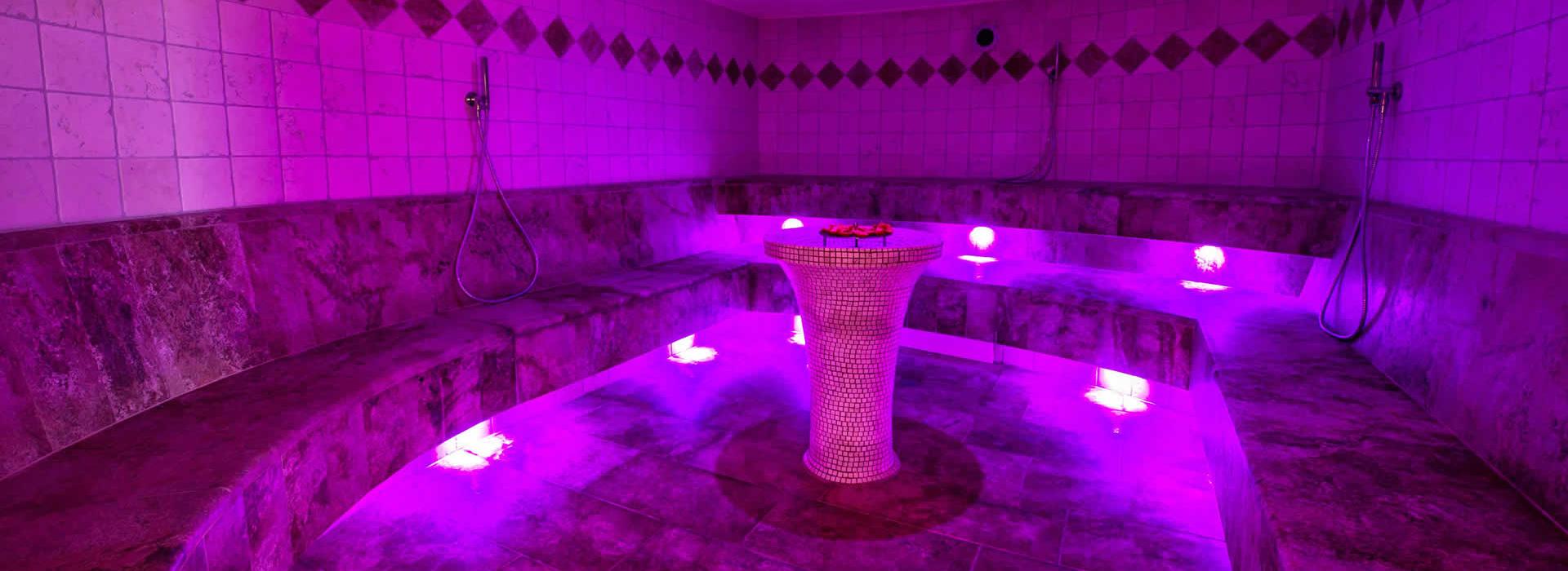 Hammam con illuminazione viola, panchine in pietra e docce a muro.