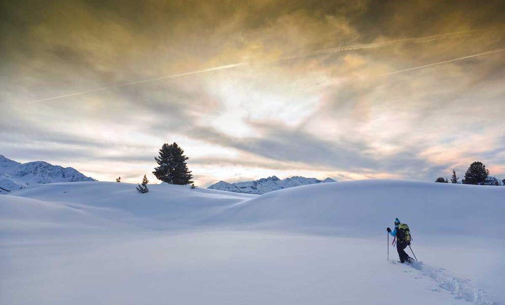 Scialpinista attraversa un paesaggio innevato al tramonto, con montagne e cielo colorato sullo sfondo.